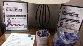 On a wooden table sits a lamp, flyers in both English and Spanish for the 'Every1 Knows Some1' Vigil event.