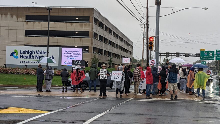 About two dozen people, many with signs like "fire Jenssen," stand in the rain in front of a Lehigh Valley Health Network sign.