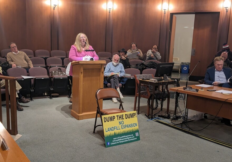 A woman speaks at a podium, with a sign in front of it reading "dump the dump"