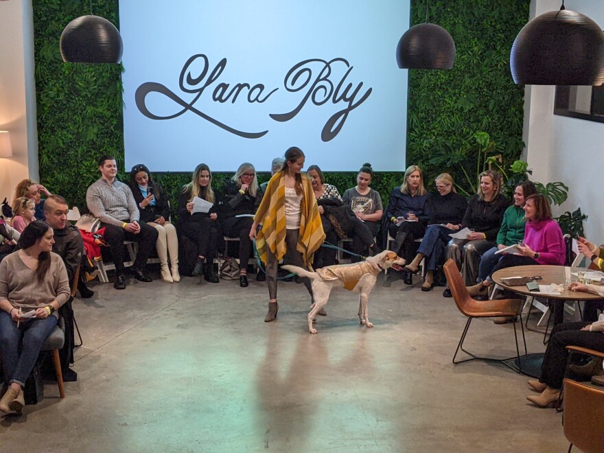 Dog mom and pup rock yellow poncho and gold-white vest. In the background in cursive font a large "Lara Bly" sign is nestled by a wall of green foliage. Attendees sit in a semi-circle  smiling at the models.