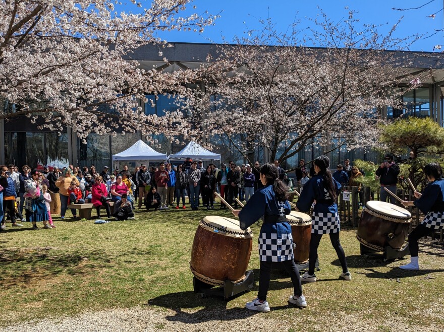 Three drummers with large wooden sticks beat on large, round wooden drums