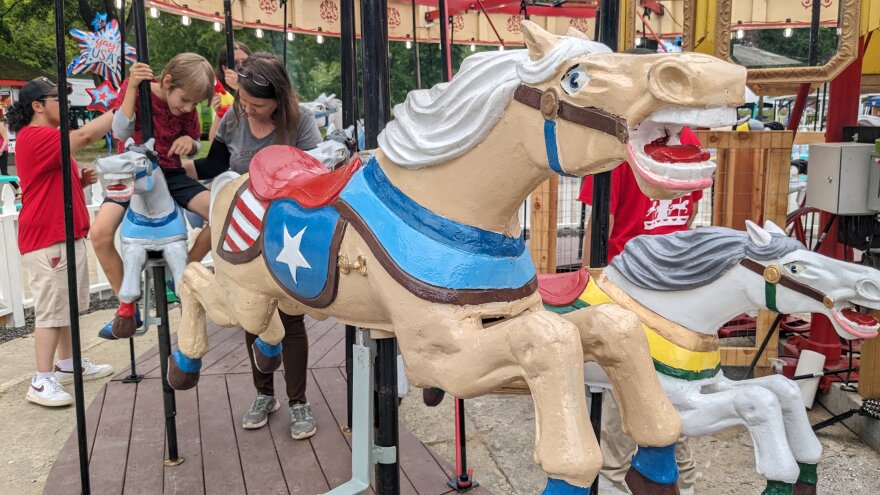 a close up of a carousel horse, with a mother and son seated on another horse in the background.
