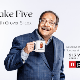 Older man in a blue blazer over a blue sweater holding a white drinking mug.