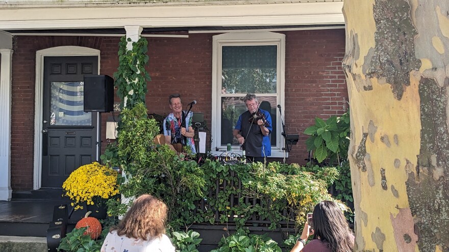 Two men, one with a guitar and one with a violin, stand in front of microphones on someone's front porch