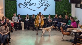Dog mom and pup rock yellow poncho and gold-white vest. In the background in cursive font a large "Lara Bly" sign is nestled by a wall of green foliage. Attendees sit in a semi-circle  smiling at the models.