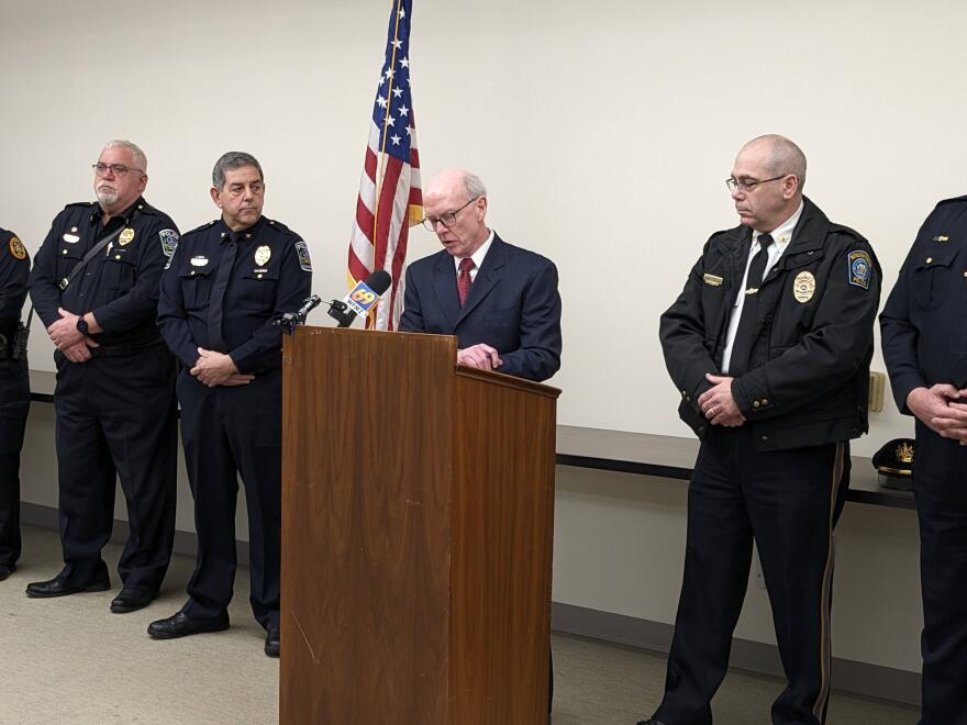 A man in a suit speaks at a podium, flanked by police officers in uniform.