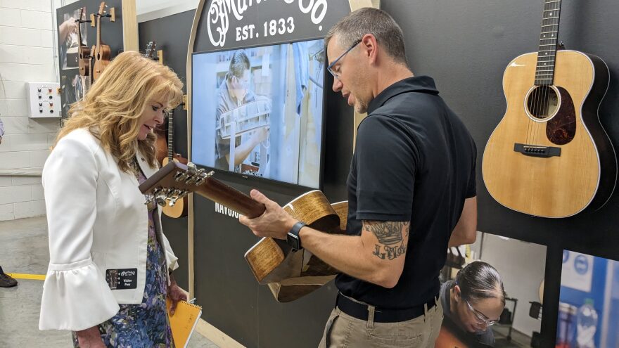 A main in a black polo shirt holds an acoustic guitar, as a woman in a white shirt takes a closes look at the guitar.