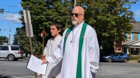 A pastor stands next to a young man with a bronze cross outside by a street