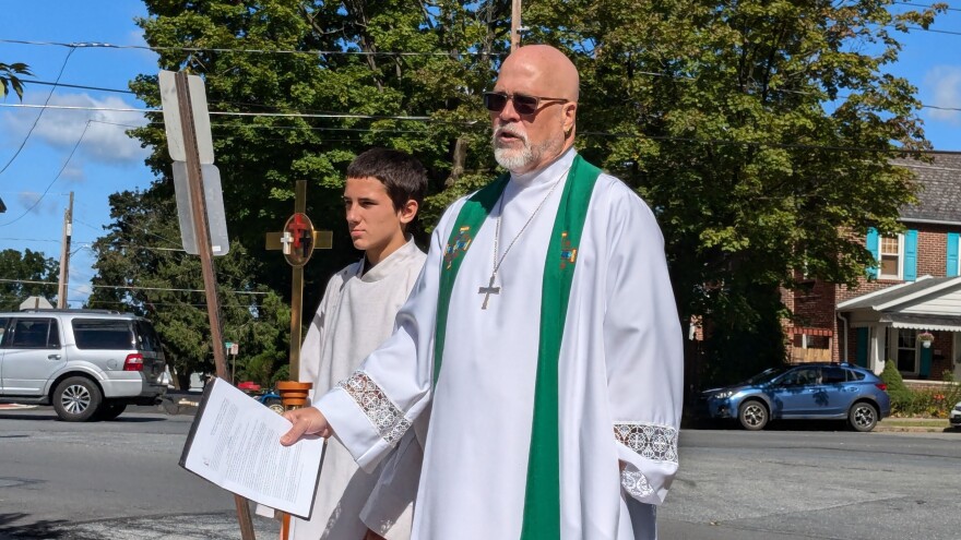 A pastor stands next to a young man with a bronze cross outside by a street