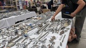 a man is standing by a row of tables covered in dozens and dozens of model and manufacturer badges off of old cars, and loads some into a plastic bin.