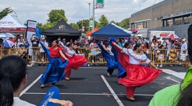 Dancers in red and blue dresses with big, flowing skirts dance in front of a crowd