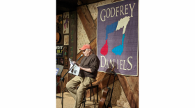 A man on a stool reads a book into a microphone
