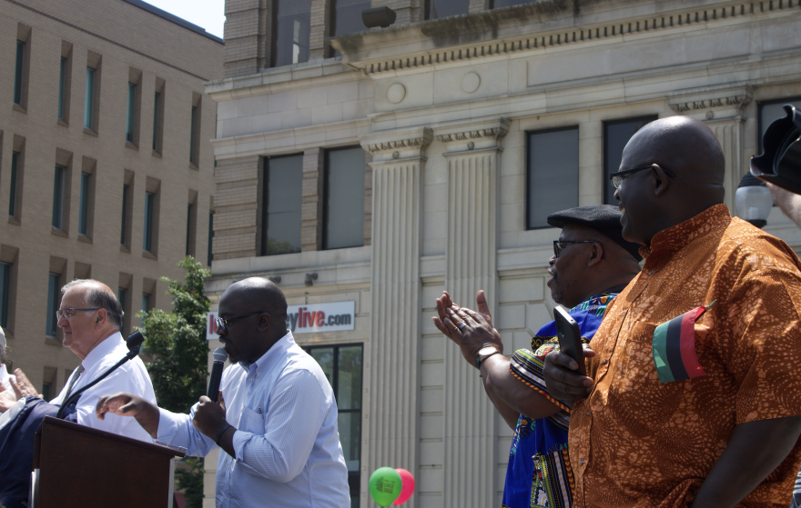 Brandon Sardik speaks from the podium at the parade. The man in the orange shirt on the right is Ray Ricketts.