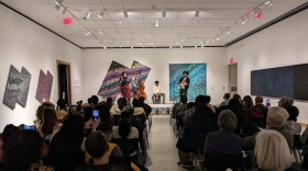 Four people, three playing instruments and one singing, stand on a stage in front of an audience. on all of the walls are large abstract paintings, each focusing on one or two different colors.