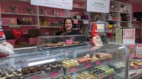 Andriana Gionis stands in behind display case full of cupcakes of different flavors like Oreo, salted caramel, and almond joy. Behind her is a sign for different cakes the shop offers along with a display of cake stands and packaging.